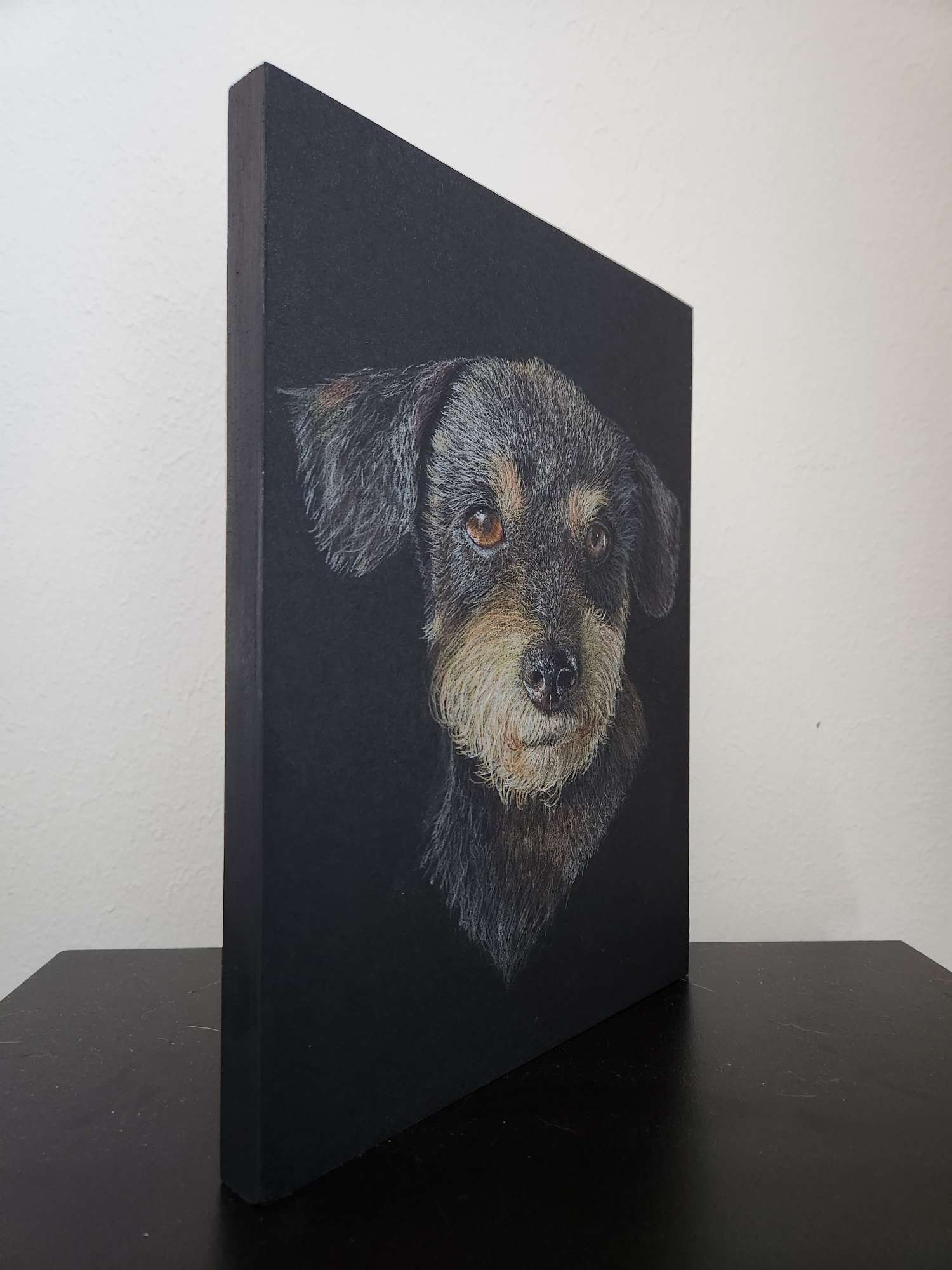 Colored pencil portrait of a young black and tan puppy drawn on black paper that's been mounted to a cradled wood panel